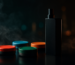 Wide dramatic image showing a vape device beside generic snus-style nicotine pouch cans with vapor swirling in a dark studio, representing snus in Bahrain and alternatives